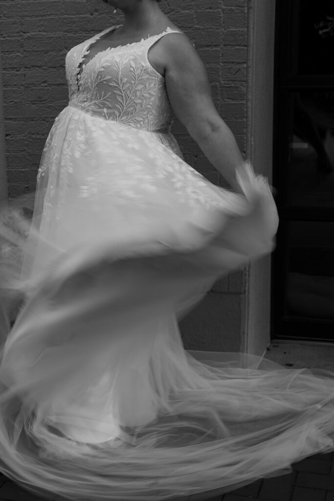 Black and white photo of a bride spinning in her dress in front of a brick wall