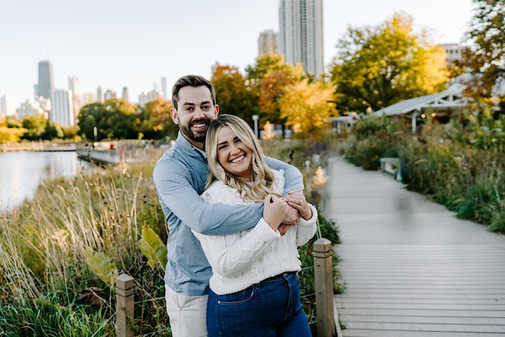 A man hugs a woman from behind as the two smile at the camera while standing on a walkway in Lincoln Park of Chicago after their proposal