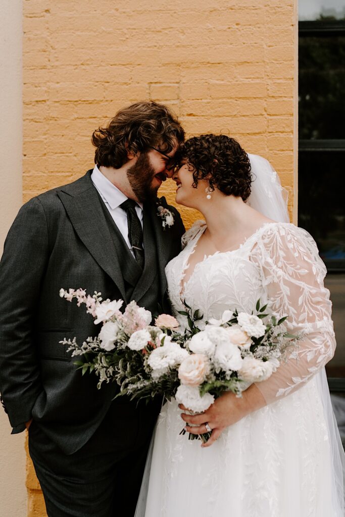 A bride and groom smile while touching noses together outside of their wedding venue while the bride holds her flower bouquet