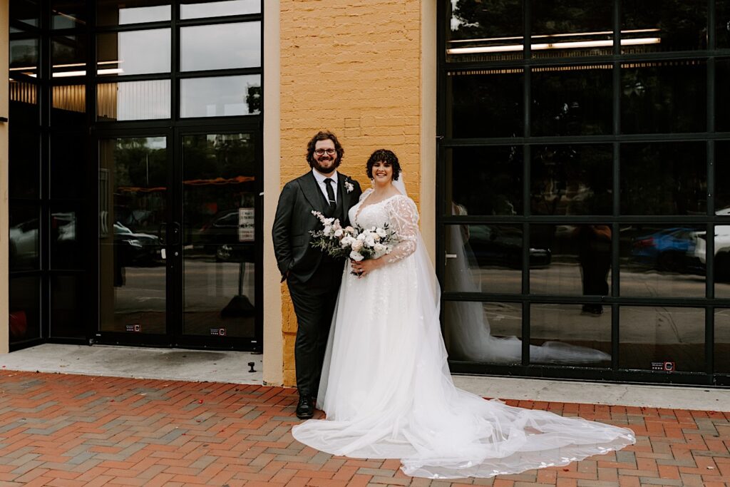 A bride and groom stand side by side and smile at the camera while outside of their wedding venue The Atrium in Milwaukee