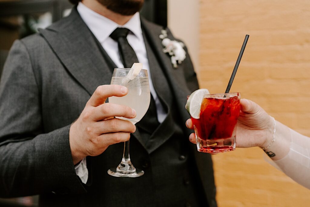 A bride and groom's hands holding drinks extend towards one another while they stand outside their wedding venue The Atrium in Milwaukee