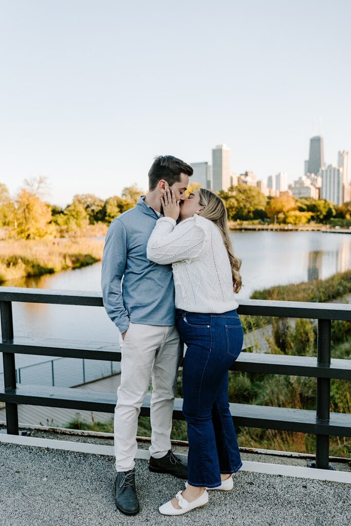 A man and woman kiss one another while standing on a bridge in Lincoln Park with a lake and the Chicago skyline behind them