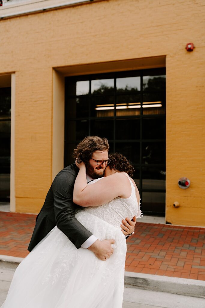A groom smiles while hugging the bride as the two stand outside their wedding venue