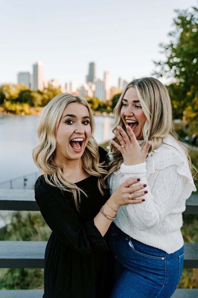A woman smiles at her friend while showing off her engagement ring, her friend is smiling at the camera while holding the other woman's arm up in the air as the two stand on a bridge in Lincoln Park of Chicago