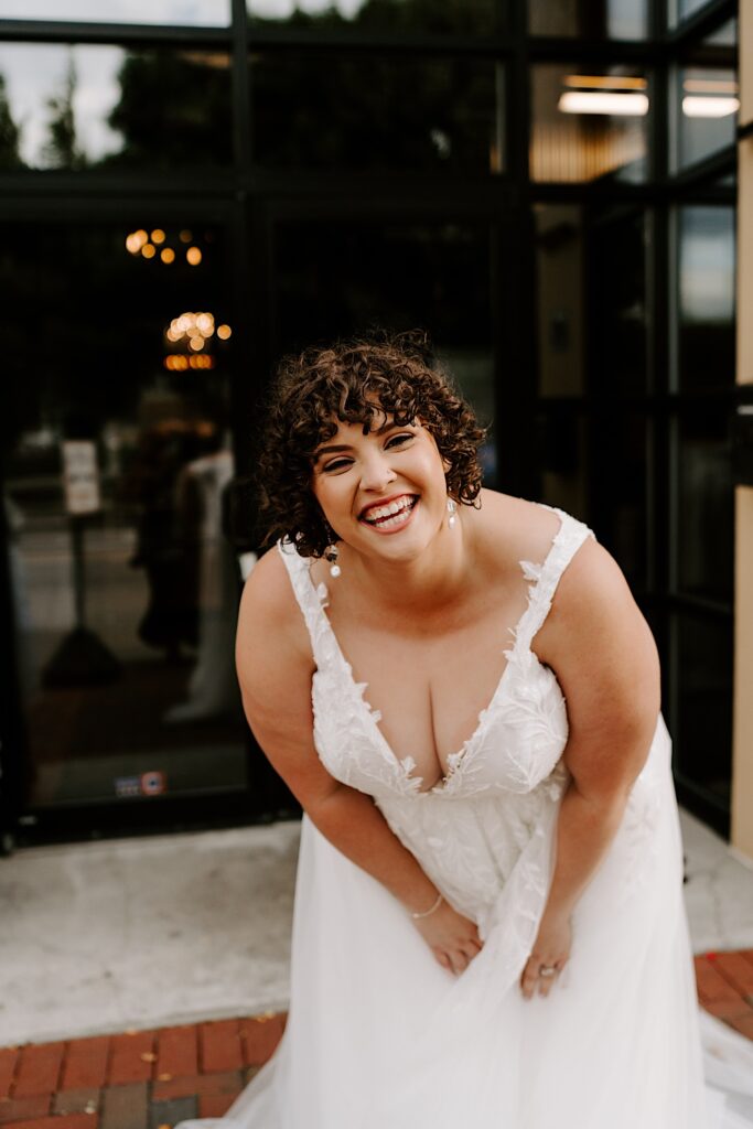 A bride leans forward and smiles at the camera while standing outside of her wedding venue