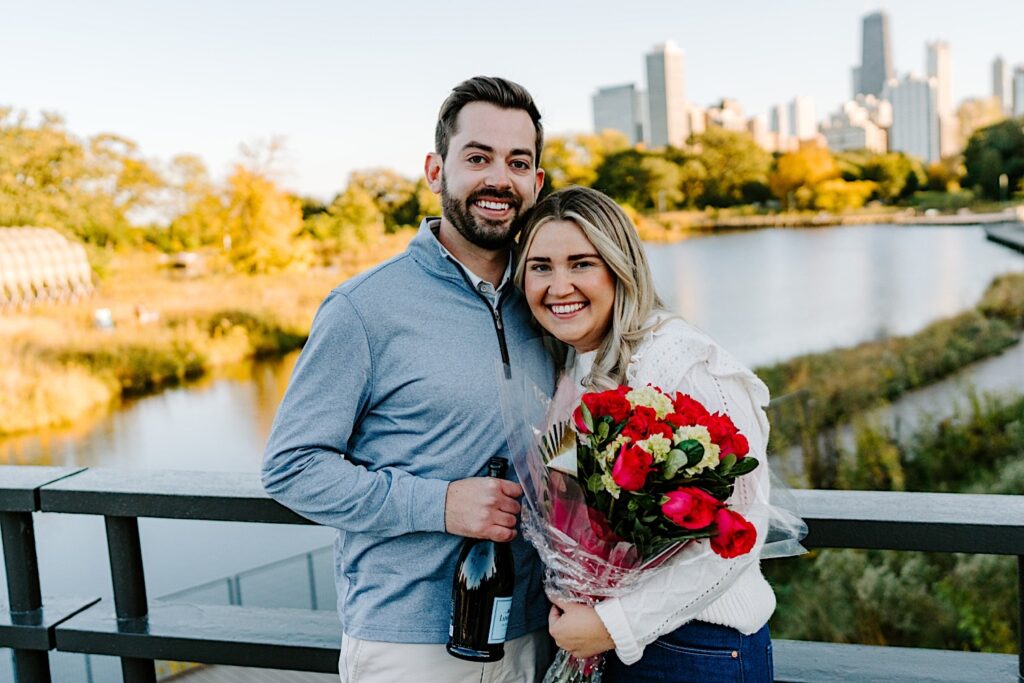 A man and woman smile at the camera while embracing as they stand on a bridge in Lincoln Park after their proposal, the man is holding a bottle of champagne while the woman holds a bouquet of flowers
