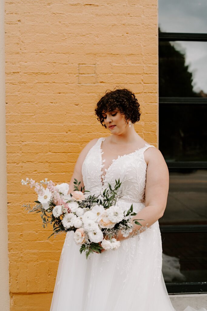 A bride stands in front of a yellow brick wall and looks down at her flower bouquet in her hand