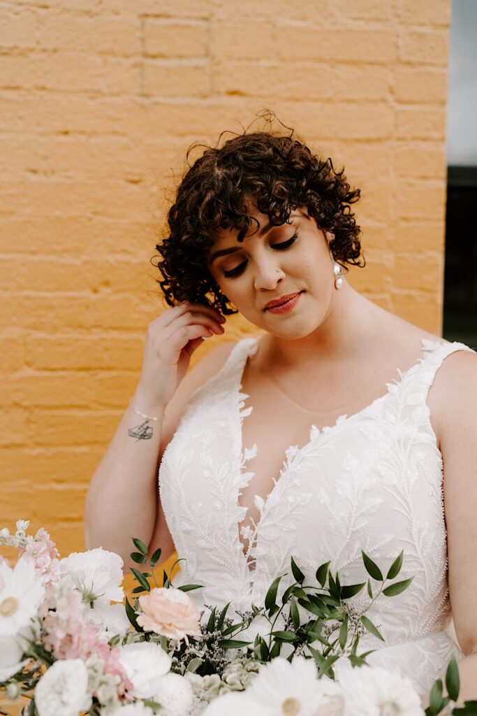A bride stands in front of a yellow brick wall and adjusts her earring with one hand while holding a flower bouquet in the other