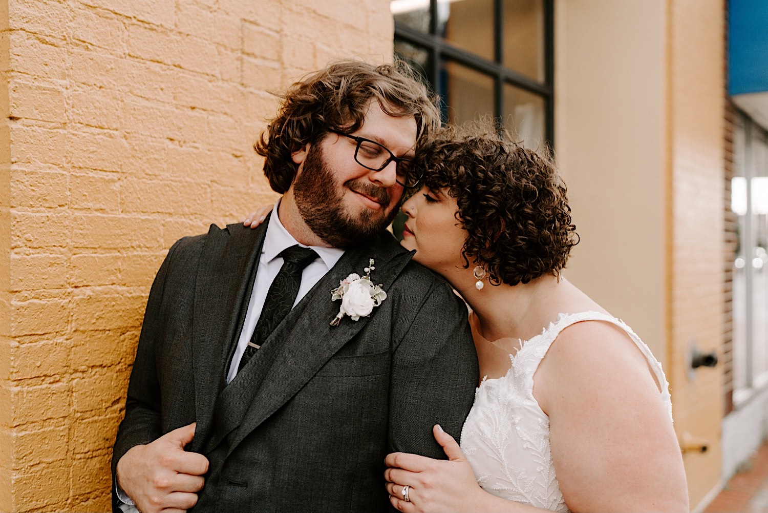 A bride leans in to kiss the groom while standing behind him as the two stand in front of a yellow brick wall outside their wedding venue The Atrium in Milwaukee