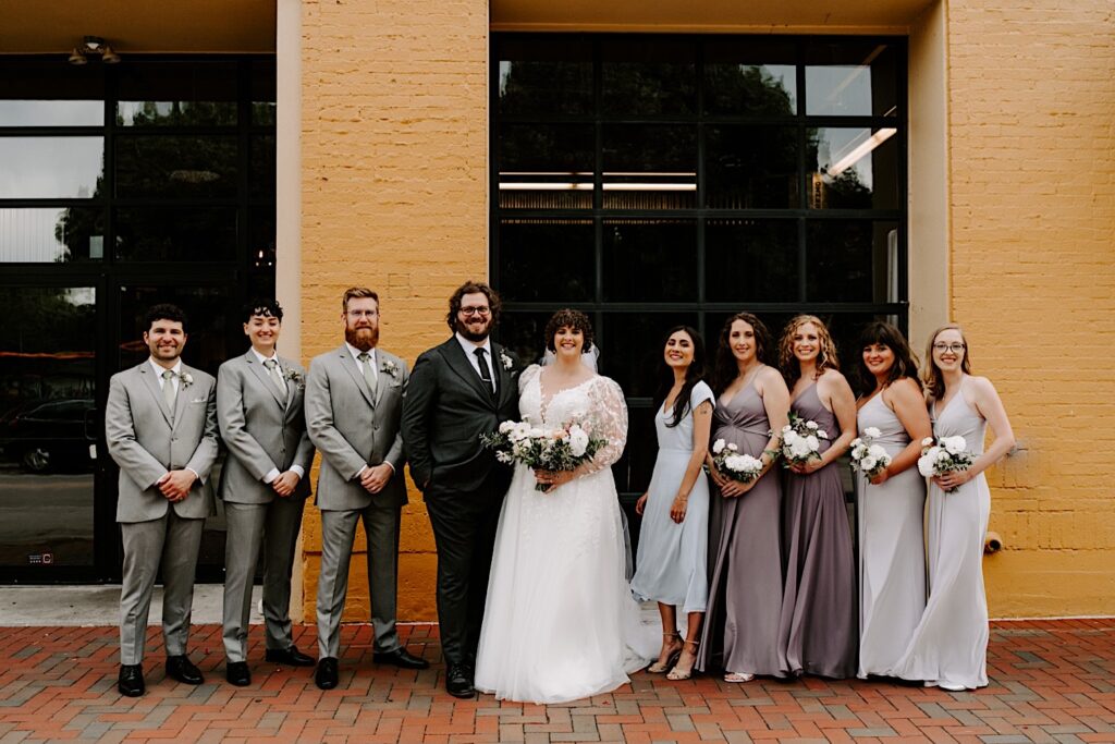 A bride and groom stand with their wedding parties on either side of them and smile at the camera together while outside their venue The Atrium in Milwaukee