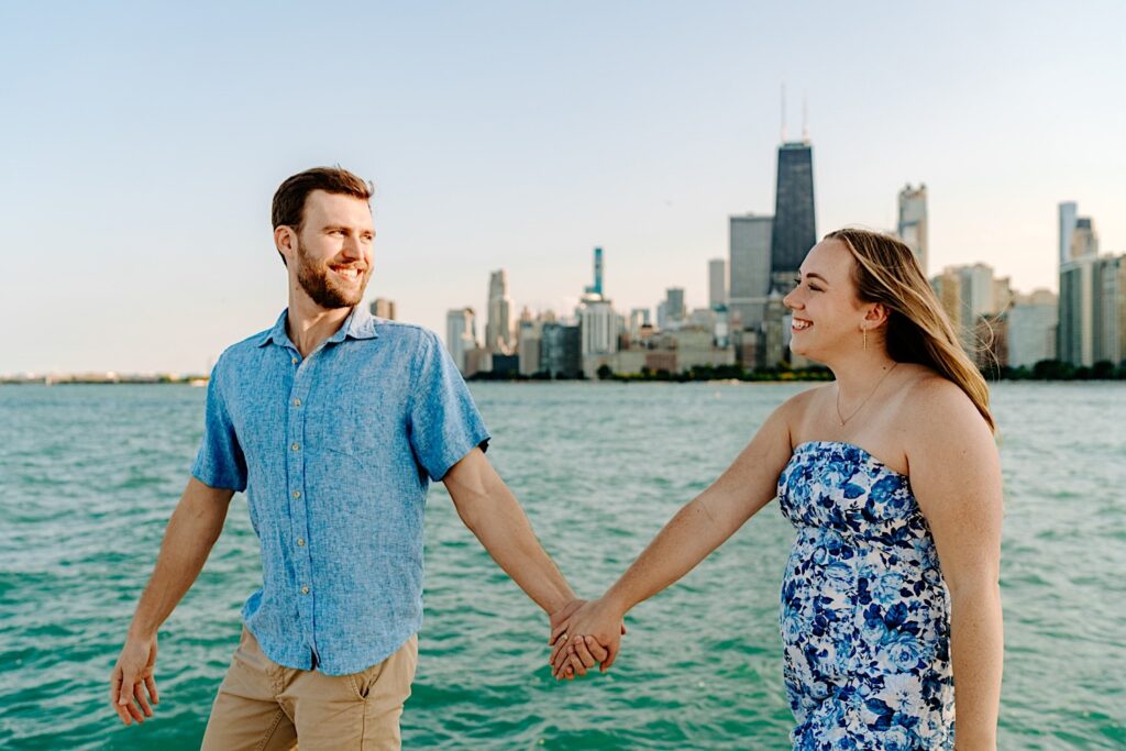 A man and woman smile at one another as they hold hands and walk along North Avenue Beach with the Chicago skyline and Lake Michigan behind them