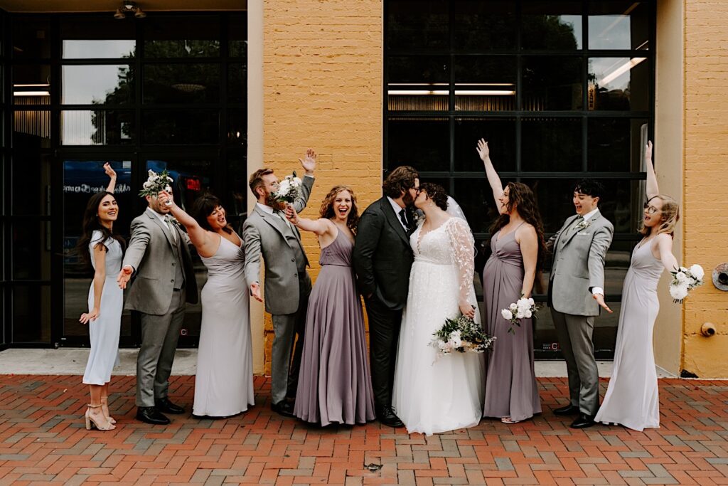 A bride and groom kiss while members of their wedding parties stand and cheer on either side of them while outside of their venue The Atrium in Milwaukee