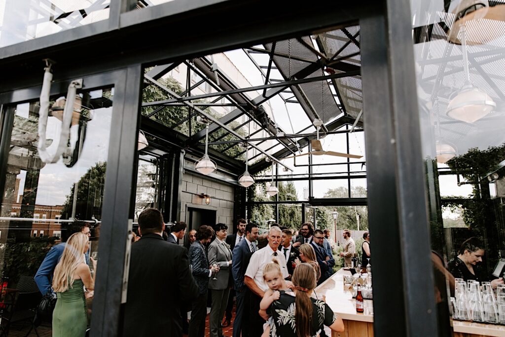 Guests of a wedding stand in a greenhouse with a bar for cocktail hour at the venue The Atrium in Milwaukee