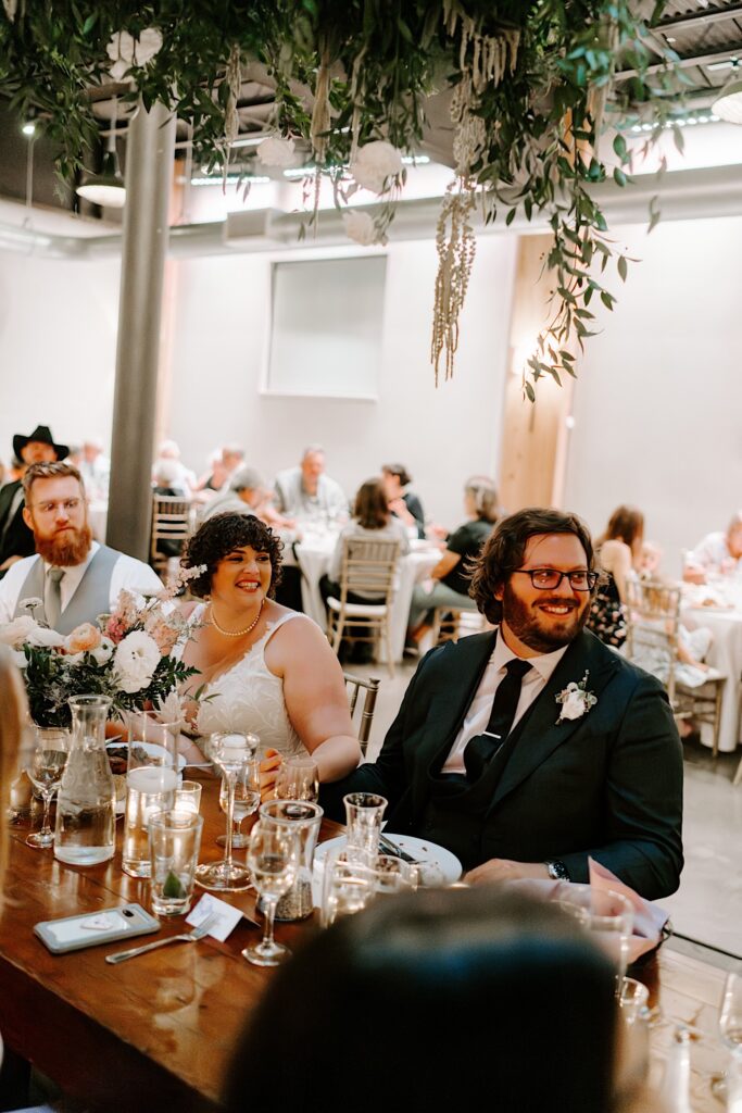 A bride and groom hold hands and smile while sitting at a table with their wedding party members during their indoor reception