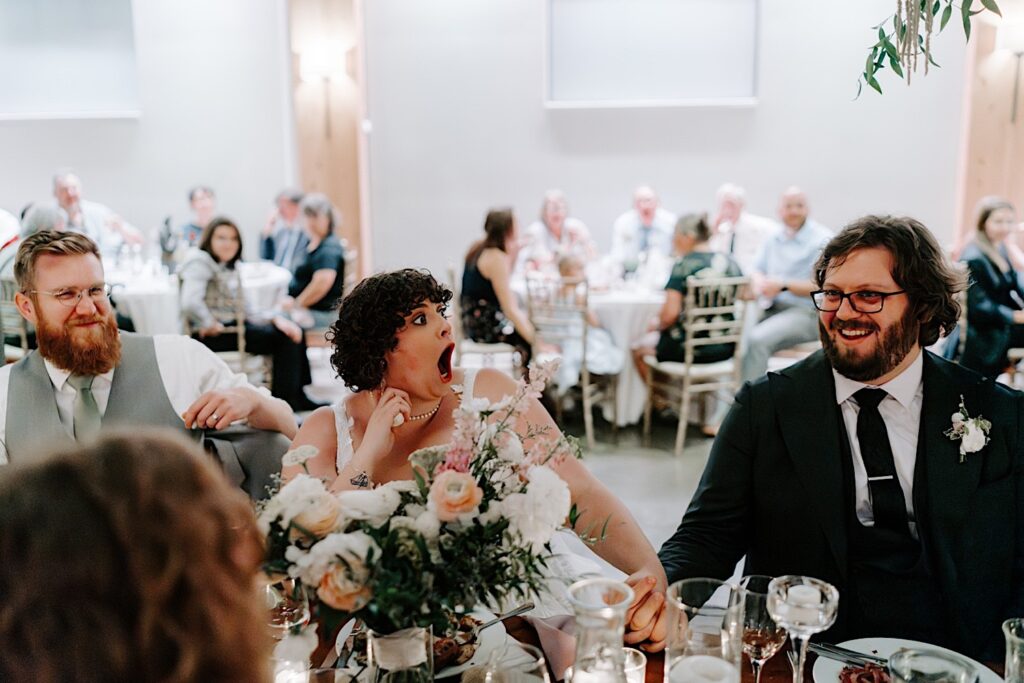 A bride makes a shocked face towards the groom as he smiles while they sit at a table during their indoor wedding reception at The Atrium in Milwaukee
