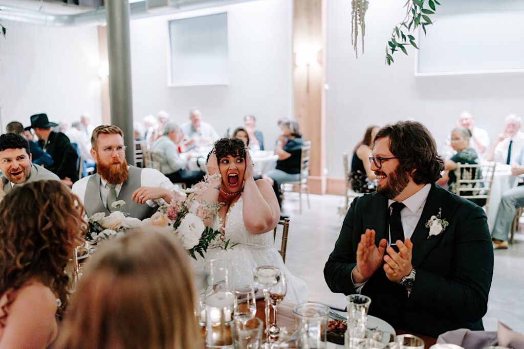 A bride makes a shocked face towards the groom as he smiles and laughs while the two sit at a table with their wedding party members during their indoor reception at The Atrium in Milwaukee