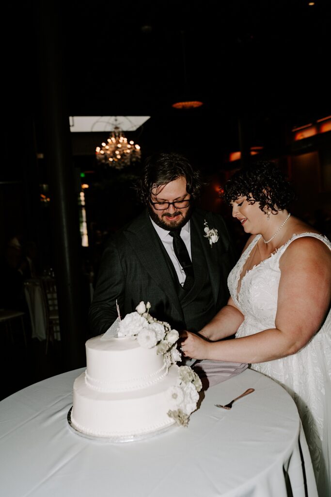 A bride and groom stand and cut their wedding cake together during their indoor wedding reception