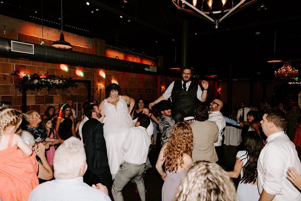 A bride and groom each sitting in a chair are lifted in the air by guests of their wedding while inside their venue The Atrium in Milwaukee