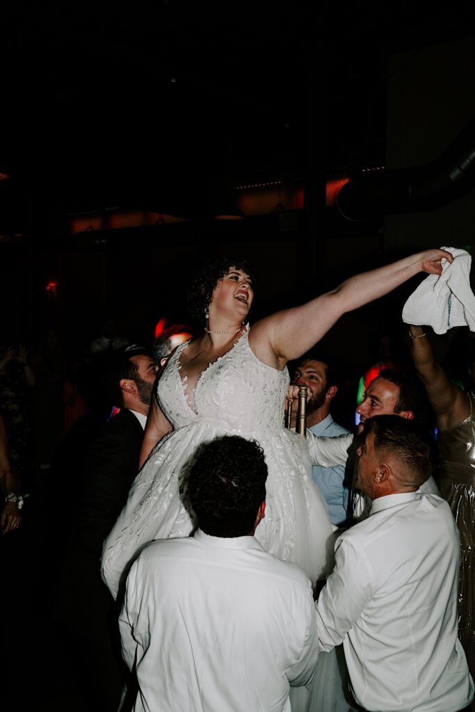 A bride sits in a chair and is lifted in the air by guests of the wedding while holding onto a white napkin