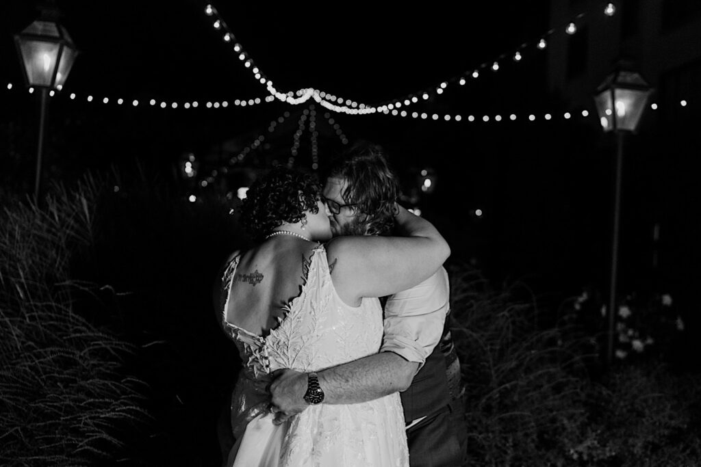 Black and white photo of a bride and groom kissing one another while under string lights outside of their wedding venue The Atrium in Milwaukee