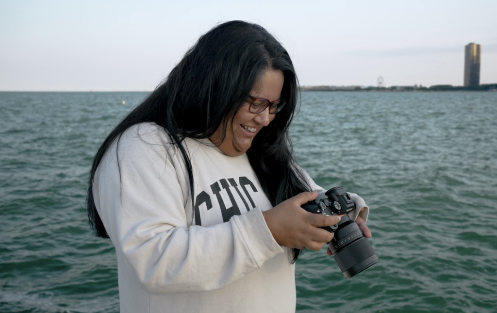 The Chicago wedding photographer, JNA Visuals, smiles as she looks down at a photo on her camera with Lake Michigan in the background