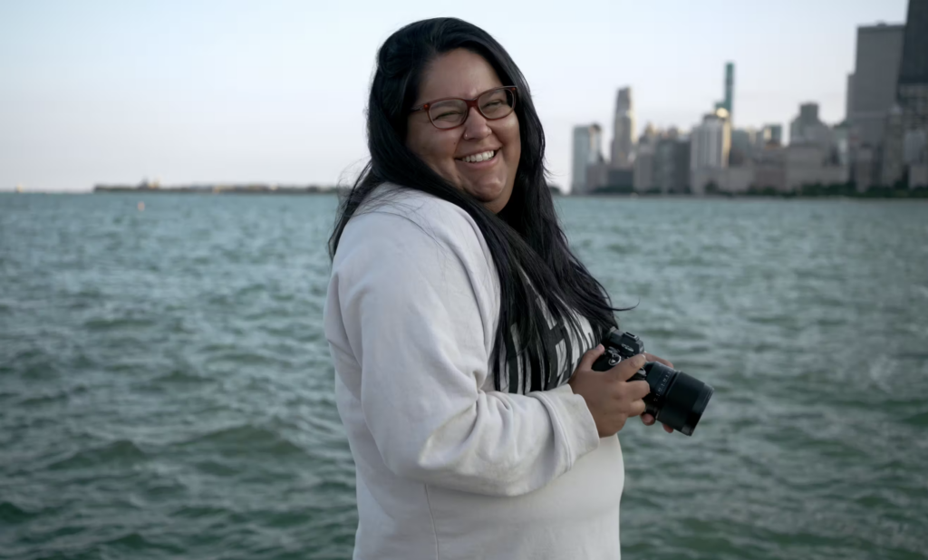 The Chicago wedding photographer, JNA Visuals, smiles at the camera while holding her camera in front of Lake Michigan and the Chicago skyline