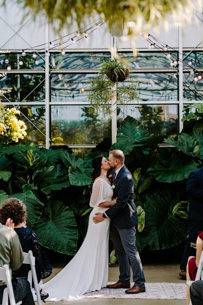 A bride and groom kiss one another inside of a greenhouse during their wedding ceremony as guests watch
