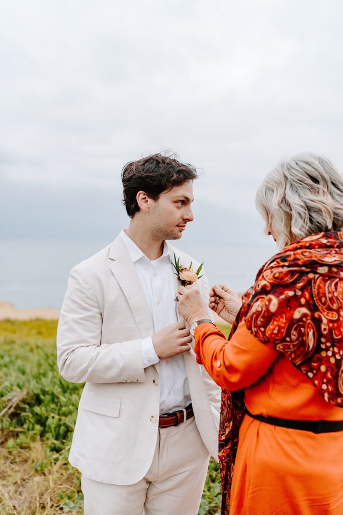 A groom looks to the side while his mother pins a flower on his suit coat on a cloudy day in San Diego
