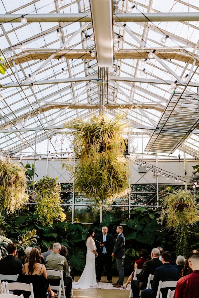 A bride and groom stand in a greenhouse during their wedding ceremony as guests are seated watching