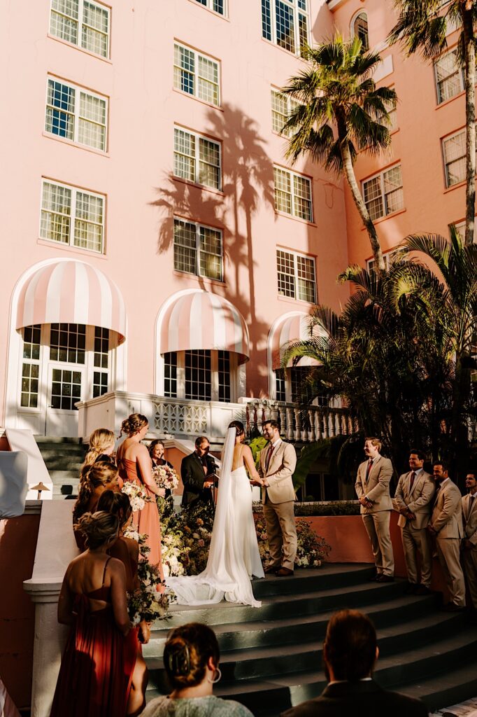 A bride and groom hold hands during their wedding ceremony on the steps leading up to a pink hotel as guests watch