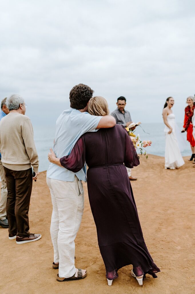 A couple hug one another while watching an elopement take place on a beach in San Diego