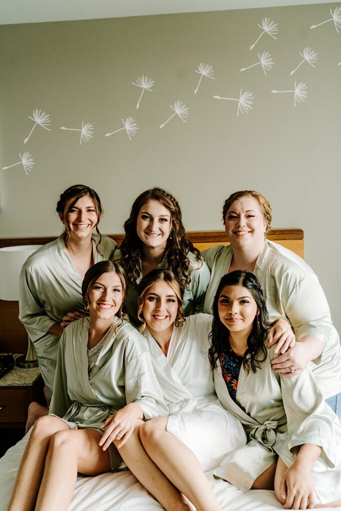 A bride sits with her 5 bridesmaids on a bed as they all smile at the camera before getting ready for the wedding day