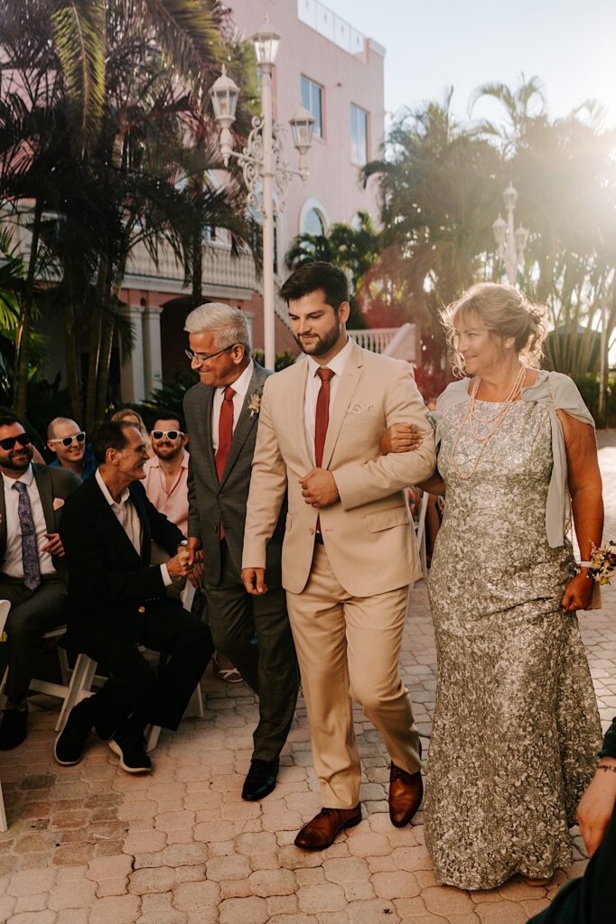 A groom is walked down the aisle by his parents with palm trees in the background
