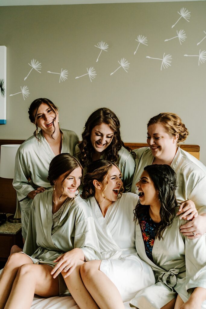 A bride sits with her 5 bridesmaids on a bed as they all smile at each other before getting ready for the wedding day