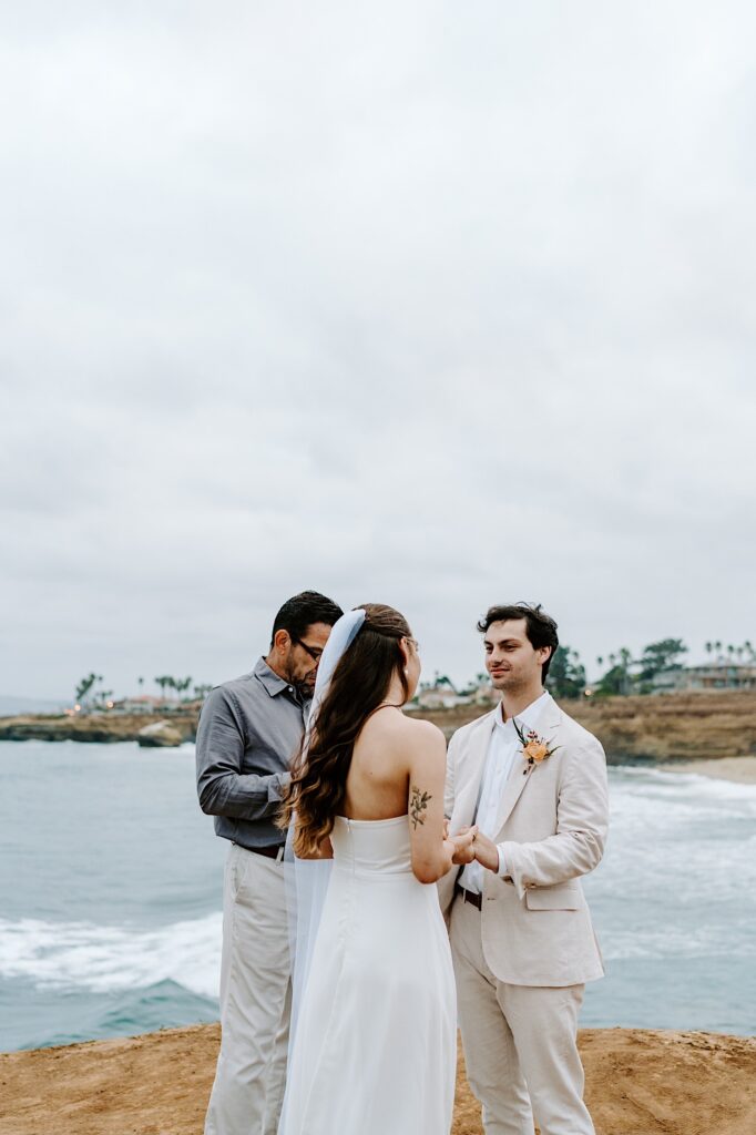 A groom smiles while holding the bride's hands during their wedding ceremony on a cloudy day in San Diego