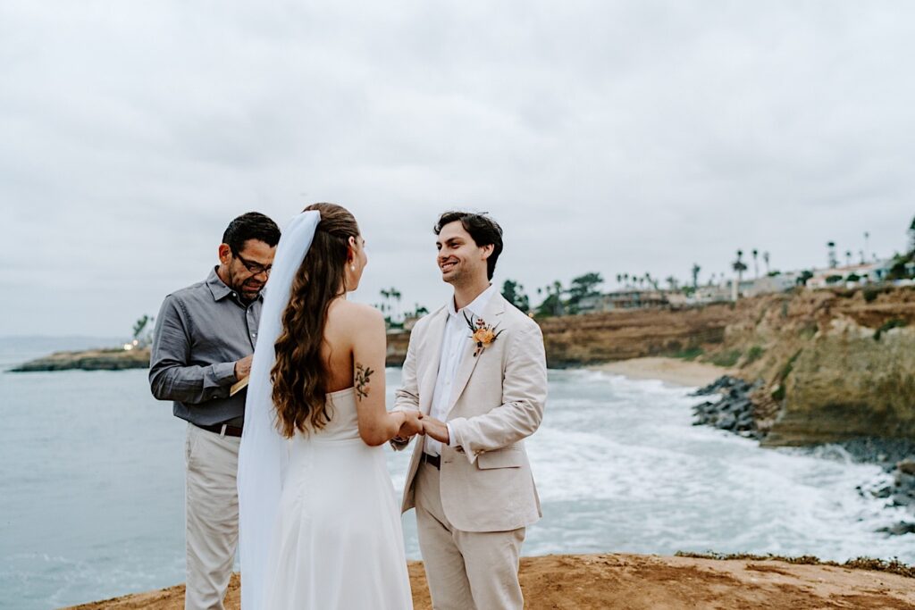 A groom smiles while holding the hands of the bride during the ceremony of their destination elopement on the cliffs in San Diego