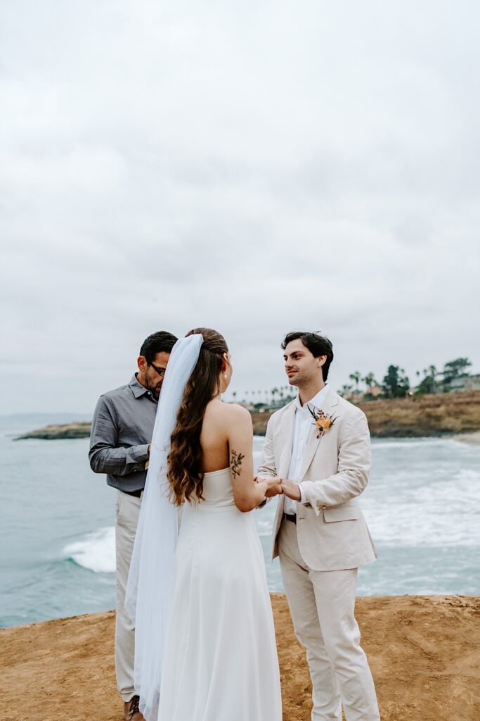 A groom holds the hands of the bride while the two stand on the cliffs in San Diego during their wedding ceremony