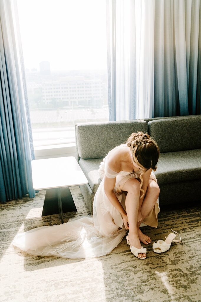 A bride sits on a couch in her wedding dress to put on her shoes in her hotel room