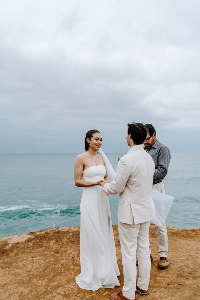 A bride smiles while holding the groom's hands during their wedding ceremony on the cliffs in San Diego