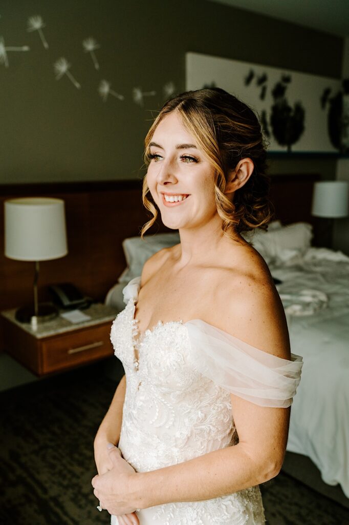 A bride in her wedding dress smiles while standing in the hotel room before her wedding