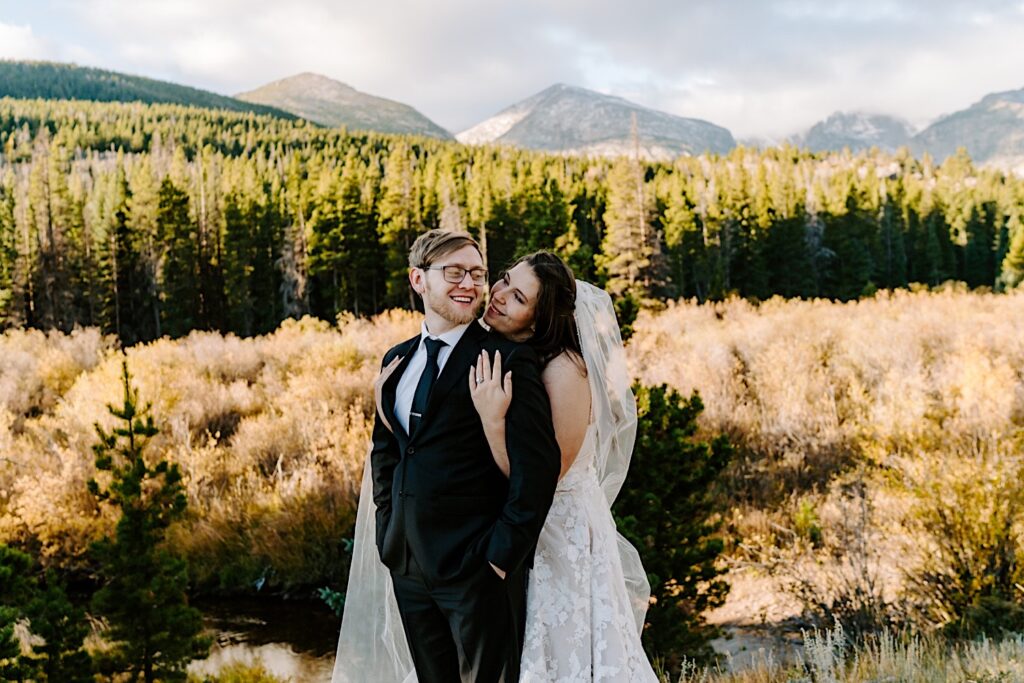 A bride hugs the groom from behind as they both smile, in the background is a forest and a mountain range, photo taken by a destination wedding photographer