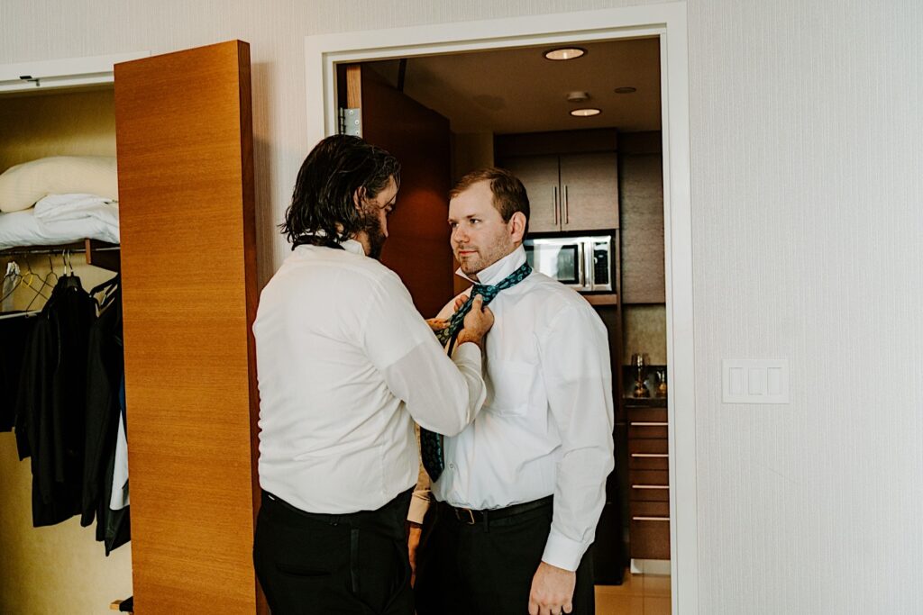 A groom smiles while a groomsman helps put his tie on for him while in their hotel room before his Chicagoland wedding