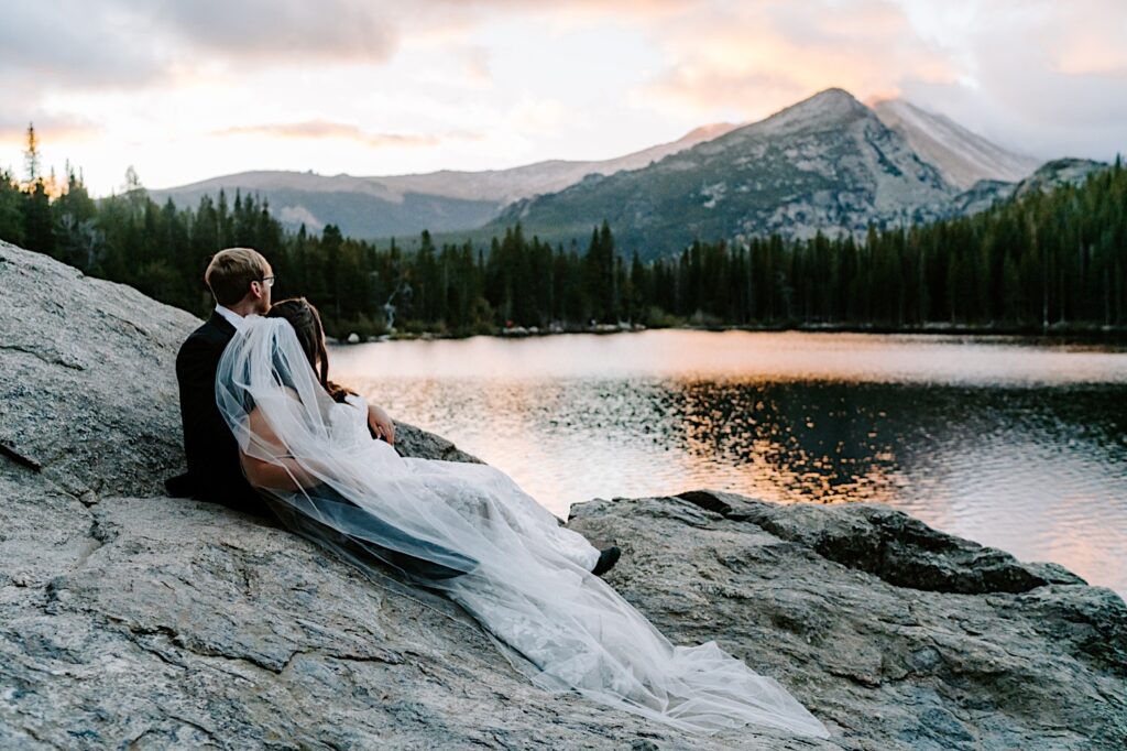 A bride and groom sit together on a rock formation looking out over a lake towards a snow covered mountain in the distance as the sun sets, photo taken by a destination wedding photographer