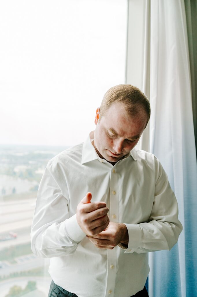 The groom of a wedding looks down as he adjusts his shirt cuff while standing in front of a window