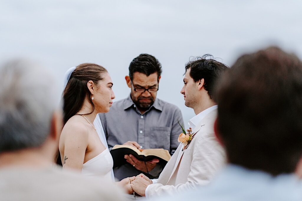 A bride and groom hold hands and look at one another while their officiant speaks and their guests watch during their destination elopement in San Diego