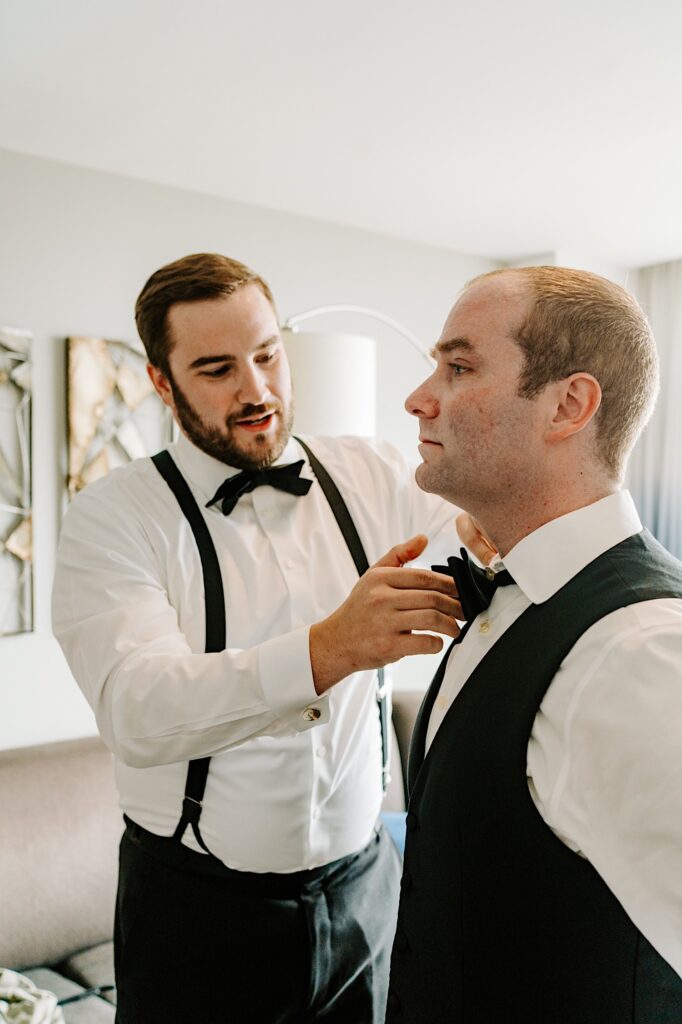 A groom stands as one of his groomsmen adjusts his bowtie while in their hotel room
