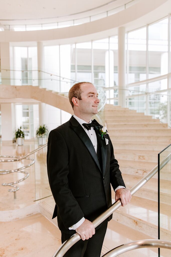 A groom stands in the lobby of a hotel in front of a large staircase and smiles as he waits for the bride