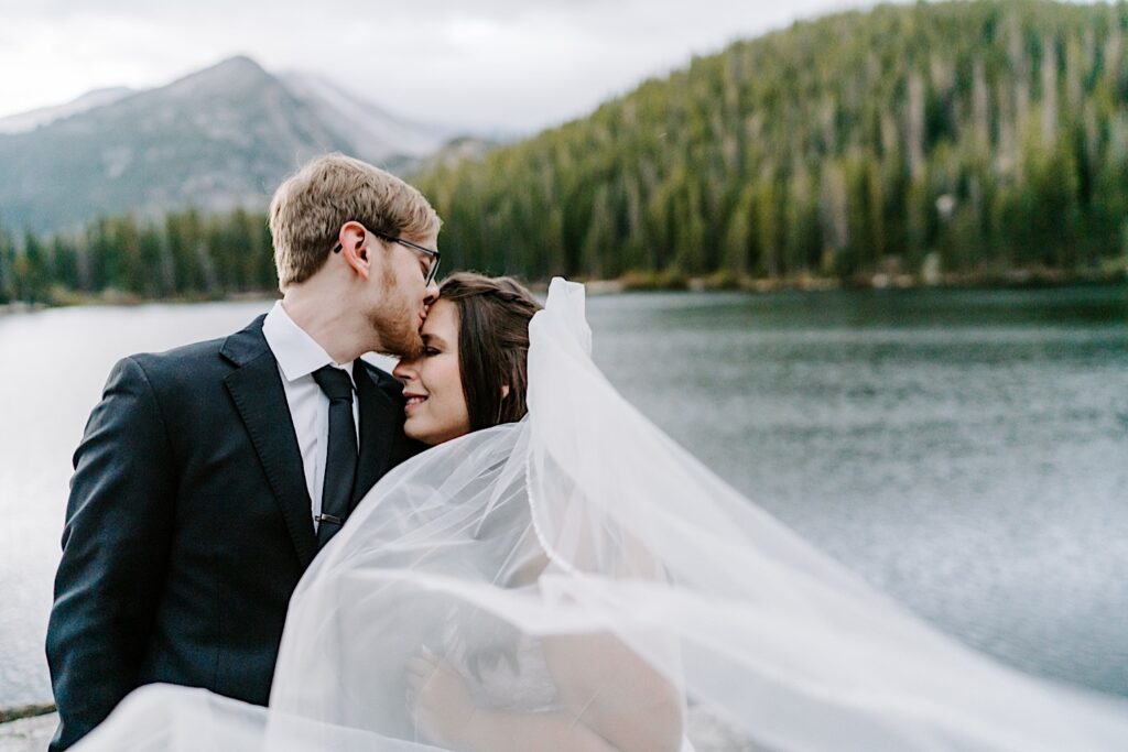 A bride smiles as the groom kisses her forehead while they stand in front of a lake with mountains in the distance behind them, photo taken by a destination wedding photographer