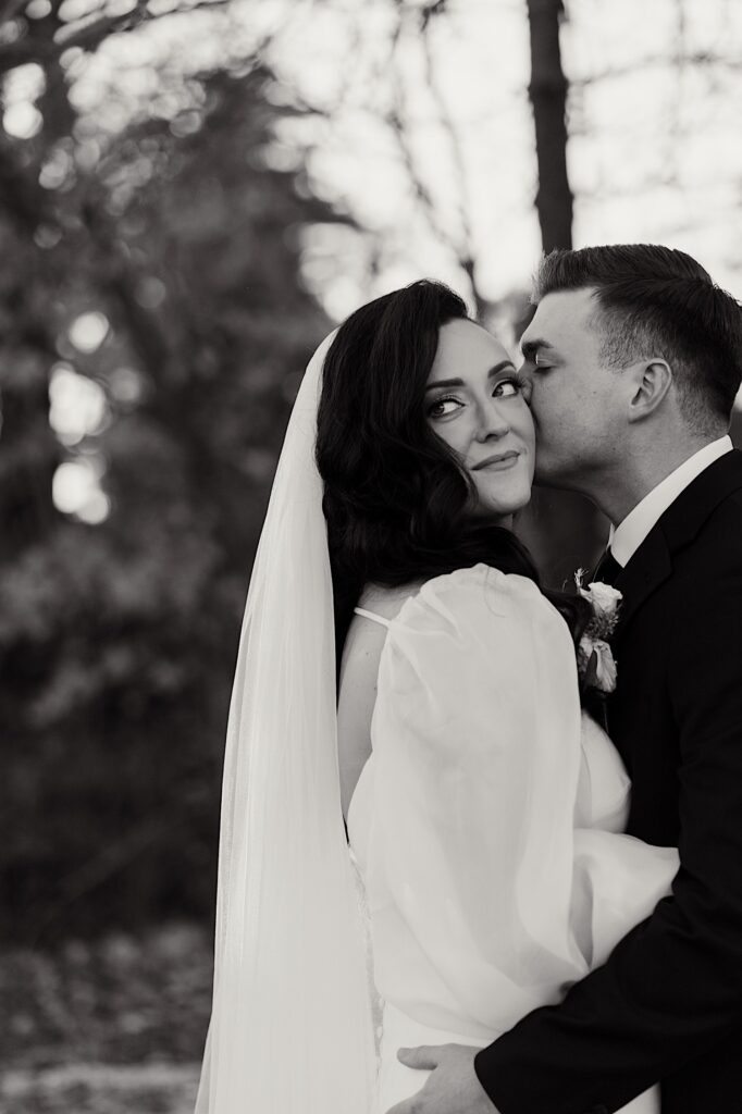 Black and white photo of a bride smiling as the groom kisses her on the cheek and hugs her