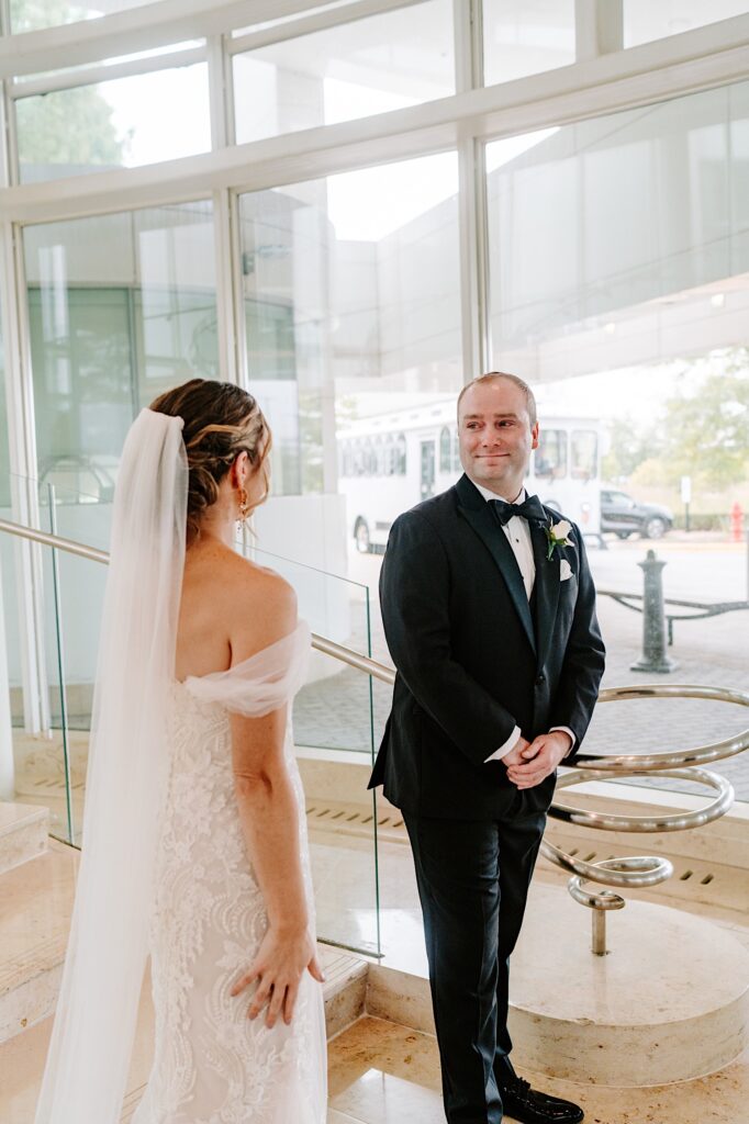 A groom smiles as he sees the bride in her wedding dress for the first time during their first look in the lobby of their hotel